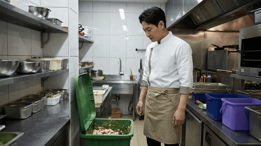 음식점 주방에서 직원이 음식물이 쌓여 있는 음식쓰레기 수거통을 바라보며 냄새와 처리 문제를 걱정하는 모습